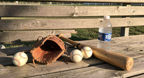 Softball gear laid out on a wooden dugout bench: a bat, a glove, two softballs and a water bottle in afternoon light.