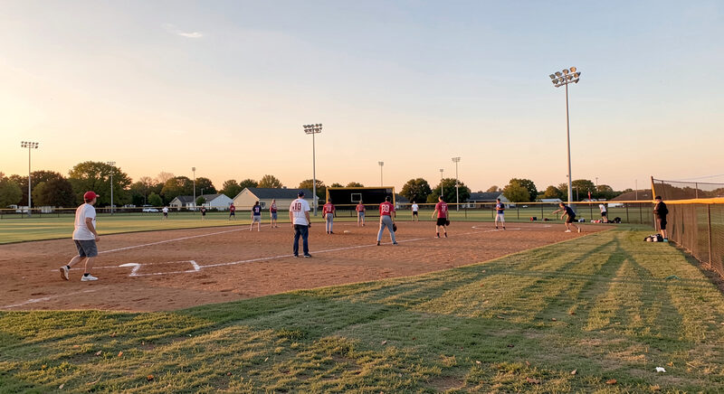 A community ballfield at golden hour with an adult softball game in progress on the infield clay, players in team shirts and a backstop under a warm sky