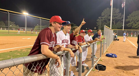 Teammates in the dugout leaning on the fence and cheering during an evening softball game under lights