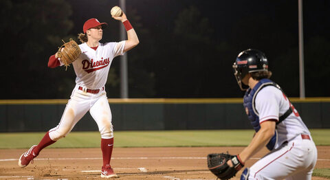 A softball pitcher wind-up under lights with the ball mid-release and a batter waiting in the box