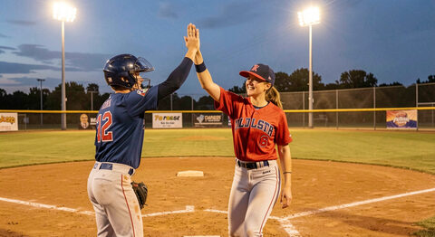 Two adult softball players high-fiving near home plate after a run scored at dusk with field lights on
