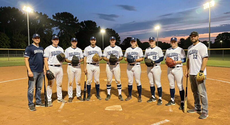 An adult co-ed softball team gathered on the infield clay at dusk under field lights, gloves and bats in hand.