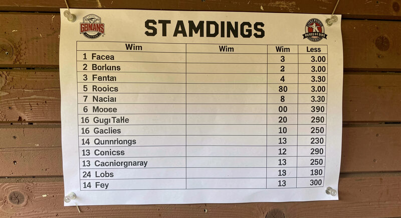 A paper standings sheet pinned to a dugout wall with team names in a column and win-loss records beside them.